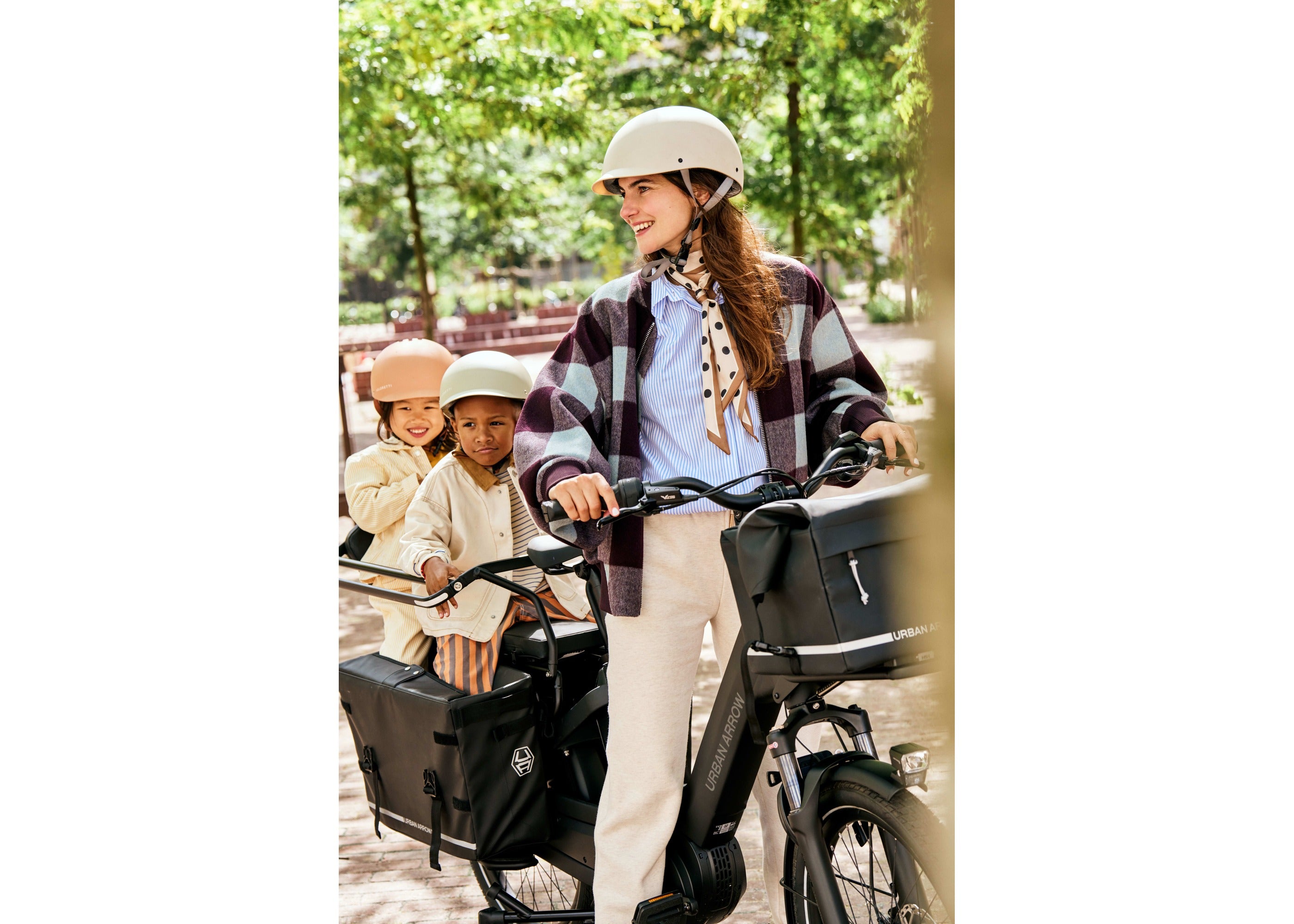 A rider pushing an Urban Arrow Breeze cargo bike with two children on board, the Front Carrier Pannier MIK loaded on the front carrier.